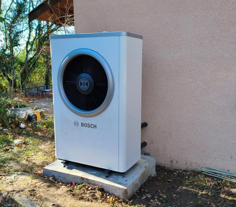 Scène extérieure avec pompe à chaleur Bosch installée sur socle béton contre mur crépi beige