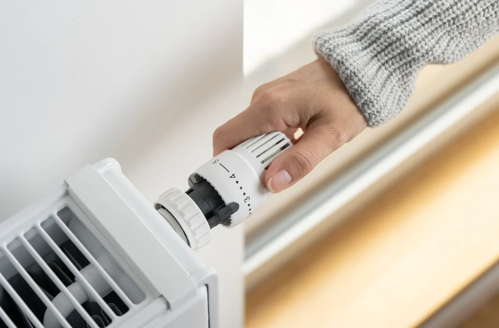 Une personne ajuste manuellement une vanne thermostatique de radiateur en intérieur, portant un pull gris