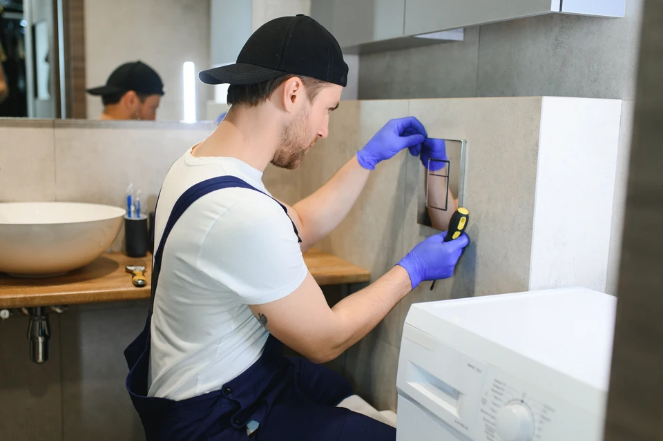 Un plombier en salopette bleue installe ou répare une douchette de douche dans une cabine blanche équipée de jets hydromassants